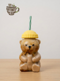 Teddy bear-shaped drink container with a yellow lid and straw on a wooden surface, branded 'Kaffeinated by Kam'.